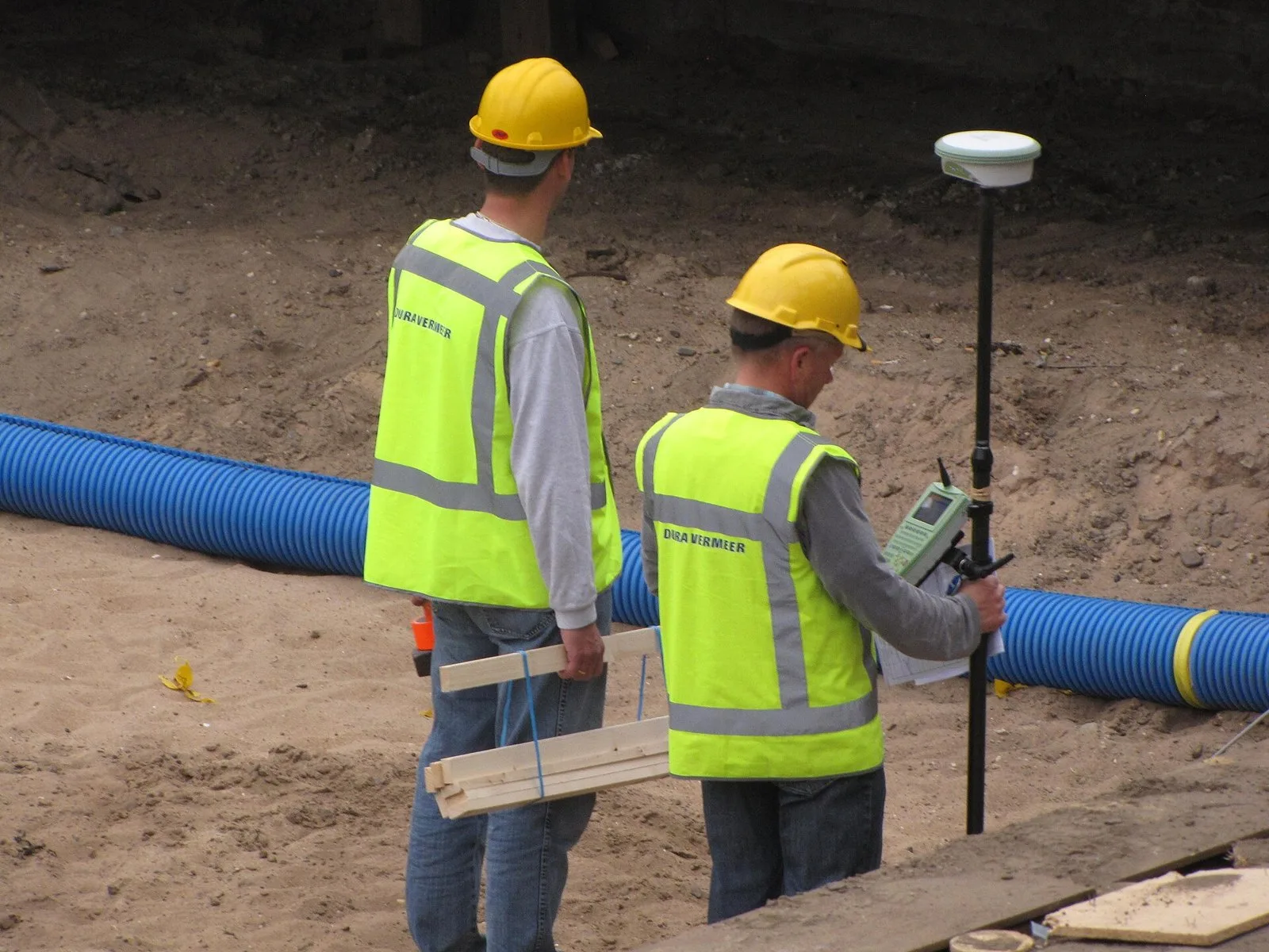 Two surveyors with a GNSS rover on a pole beside a large pipe in an excavation